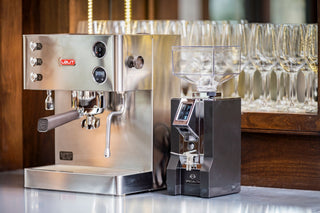 Stainless Lelit espresso machine with a portafilter on a counter and a grinder with a clear hopper in the background
