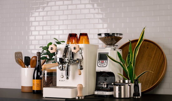 White espresso machine with a clear bean hopper beside a compact grinder on a dark counter and a plant nearby