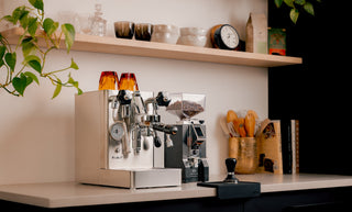 Lever espresso machine with wooden accents and two orange cups on top beside a grinder and shelves