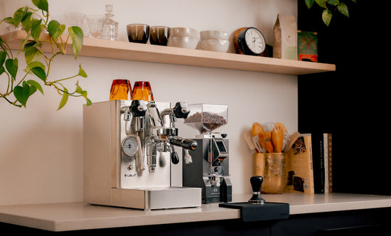 Lever espresso machine with wooden accents and two orange cups on top beside a grinder and shelves