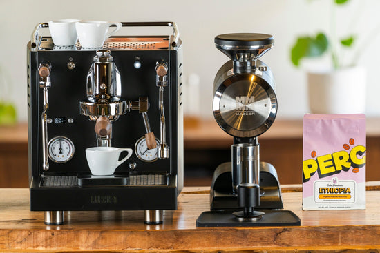 LUCCA espresso machine with two white cups on top beside a Mazzer grinder on a wooden counter