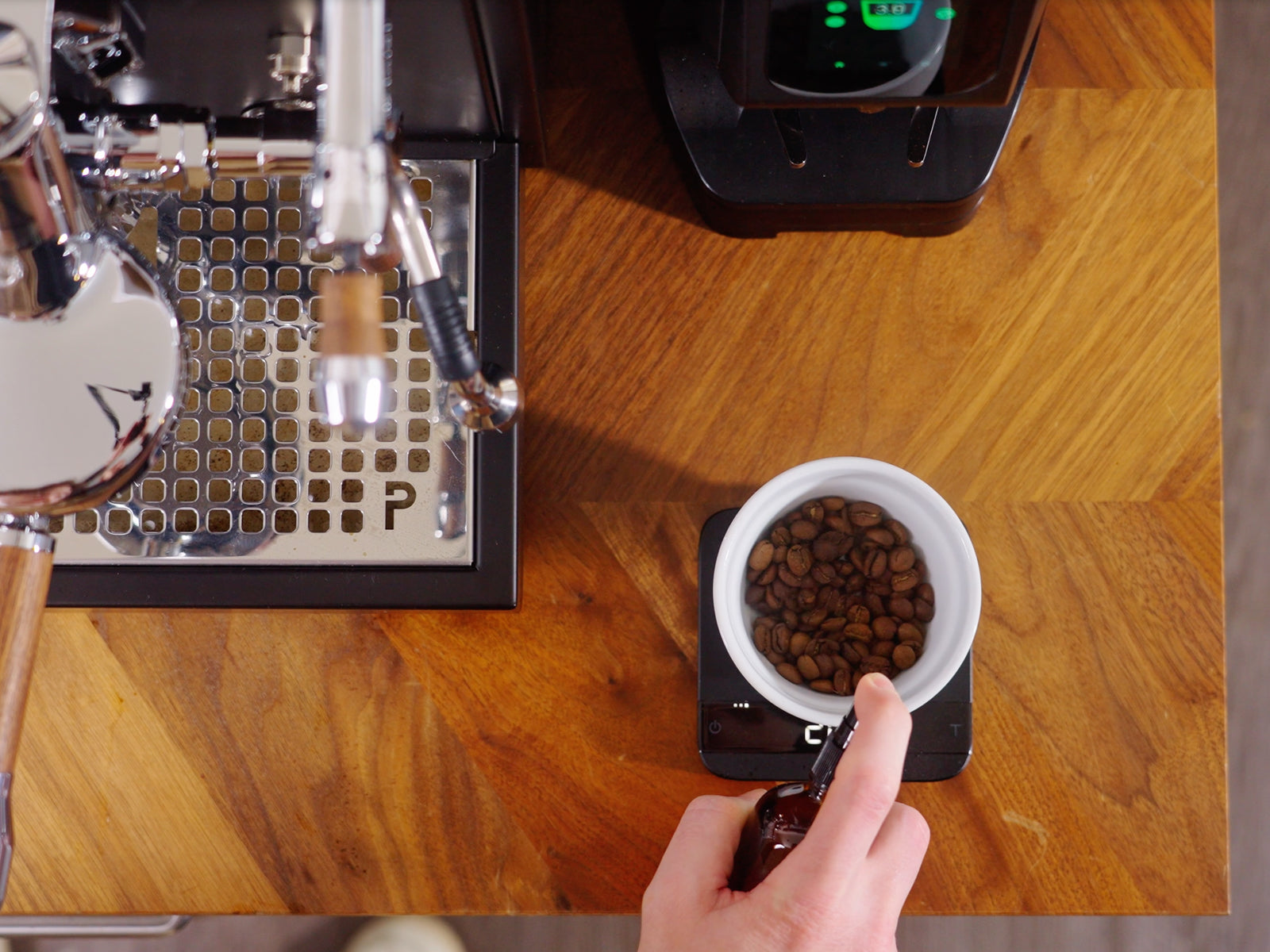 Overhead shot of a puck prep workflow with a hand pouring from a bottle into a scale beside a bowl of roasted beans.