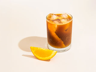 Tall glass of iced espresso tonic with ice and orange peel, accompanied by a fresh orange wedge on a pale surface