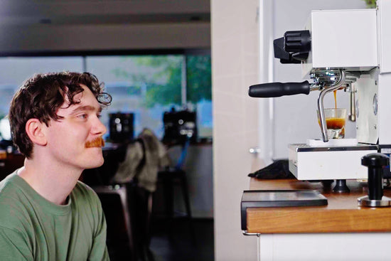 Man watches as espresso pours from a portafilter into a small glass beneath a white espresso machine.