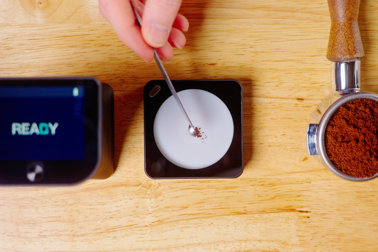 Overhead view of a digital scale with a spoon scooping coffee grounds into a white disc beside a grinder of grounds