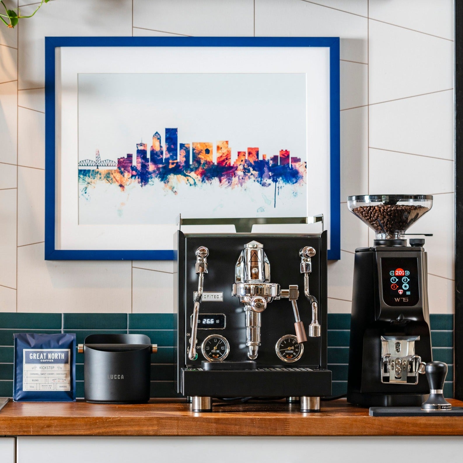 Two-group espresso machine on a wooden counter with portafilters and a knock box nearby coffee bag and kettle in view.