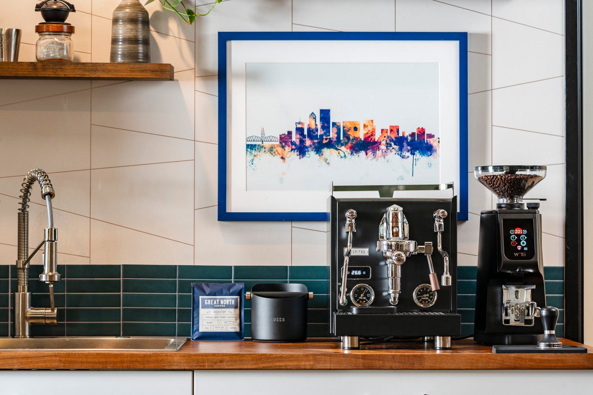 Chrome espresso machine and bean grinder on a wooden counter under a blue framed city skyline artwork