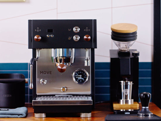 Black espresso machine with copper knobs and multiple dials on a wooden counter, a grinder nearby.