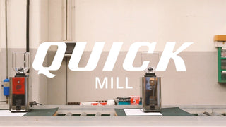Industrial coffee workshop with two espresso grinders and a large Quick Mill logo overlay.
