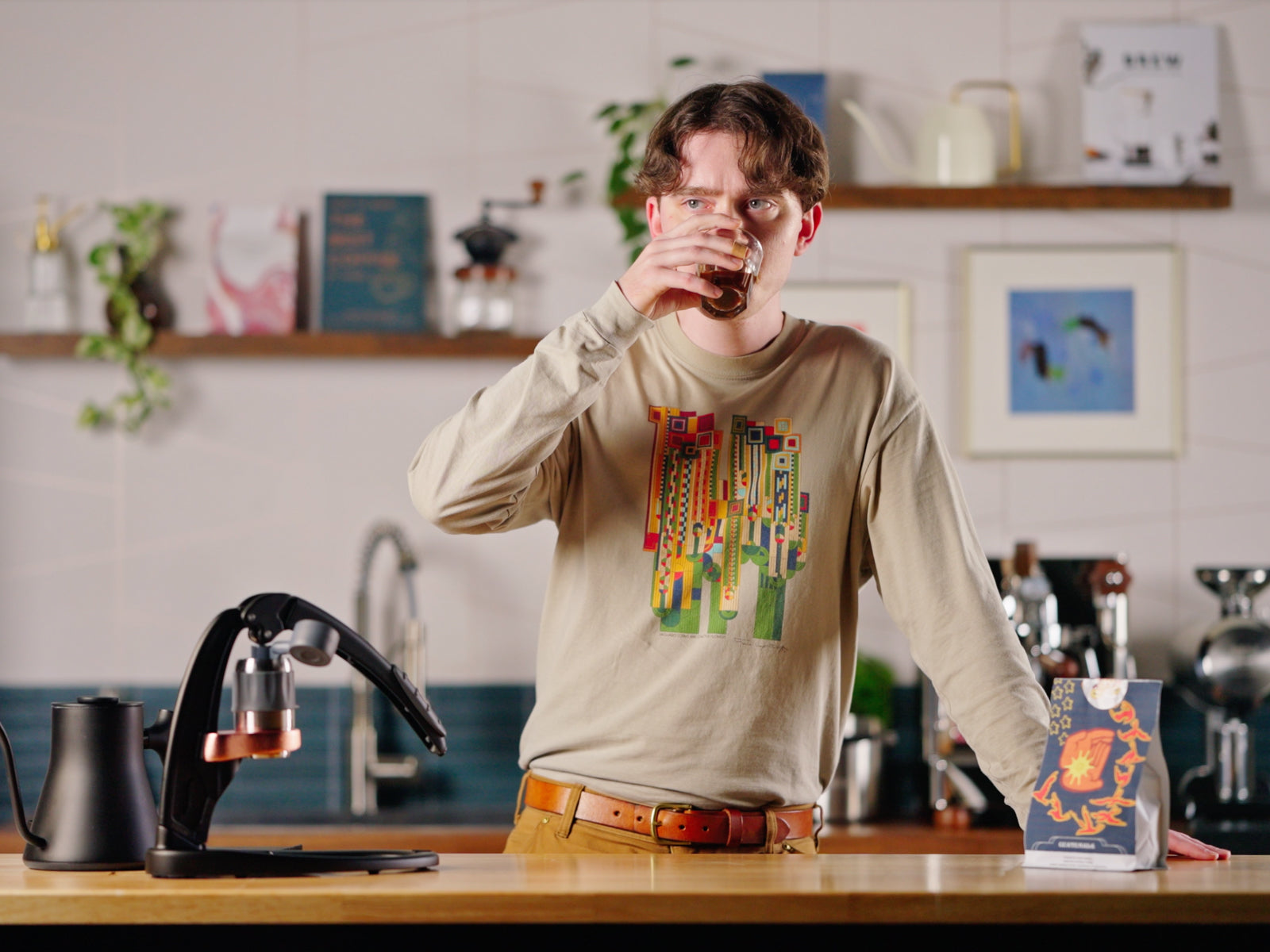 Man in a beige graphic shirt drinks from a glass at a cafe counter with an espresso machine and festive greenery.