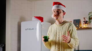 Man in a cream knit sweater wearing a Santa hat holds a green beverage in a bright kitchen.