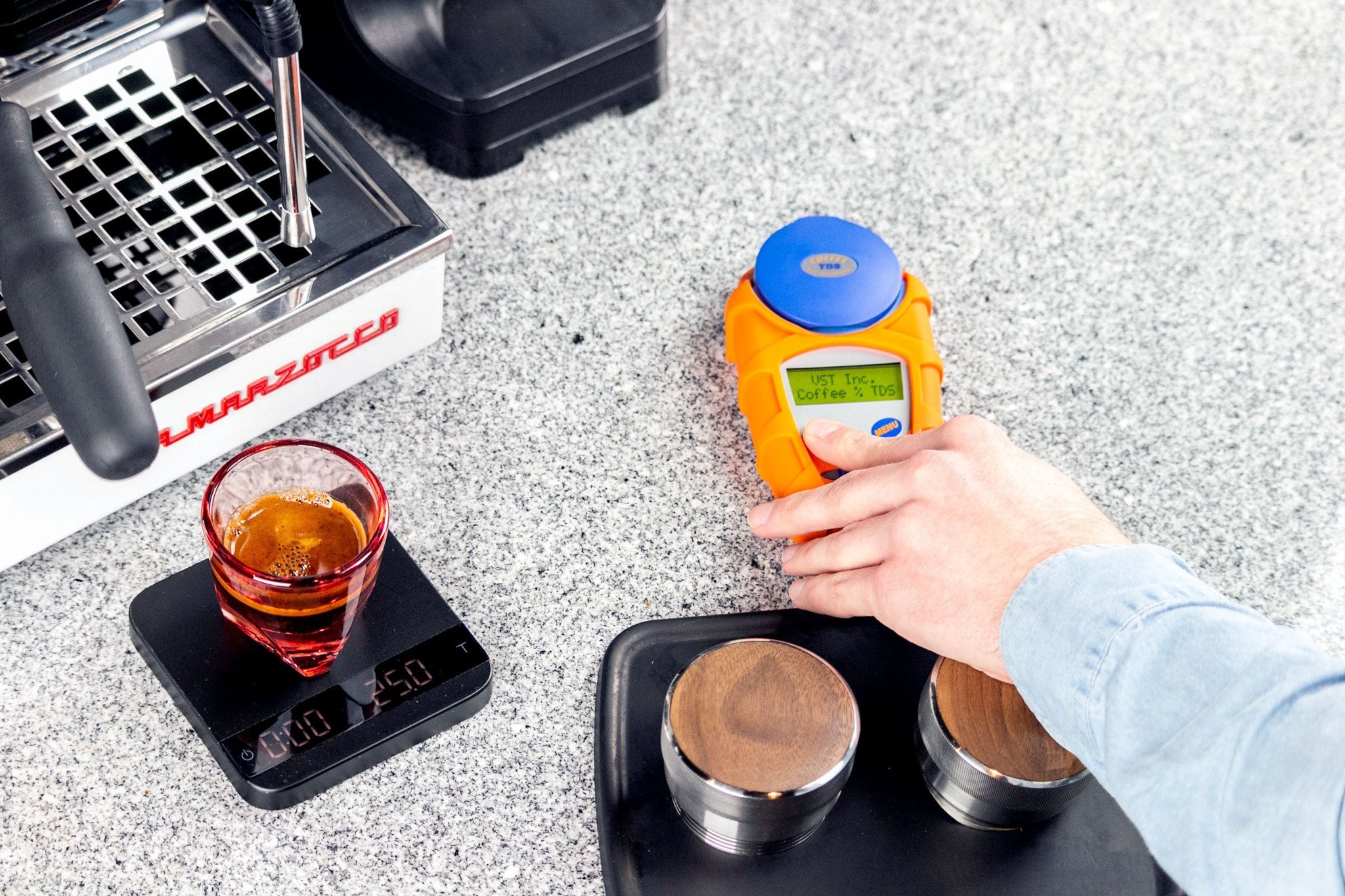 Barista uses an orange handheld coffee refractometer to measure espresso extraction beside a scale and glass of espresso.