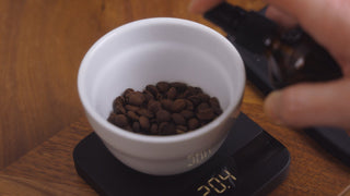 White ceramic cup filled with roasted coffee beans sits on a digital scale on a wooden counter with a hand in the background
