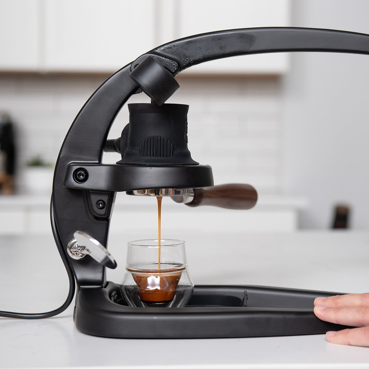 Close up of a black lever espresso machine dispensing espresso into a glass on a kitchen counter.