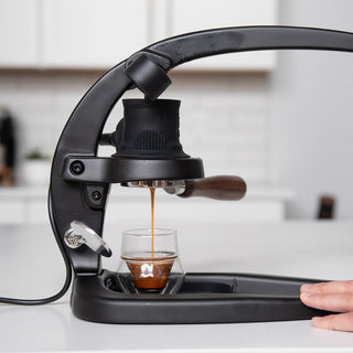 Close up of a black lever espresso machine dispensing espresso into a glass on a kitchen counter.