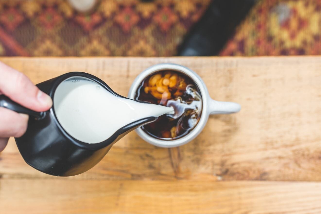 Milk pours from a black creamer into a cup of coffee on a wooden table