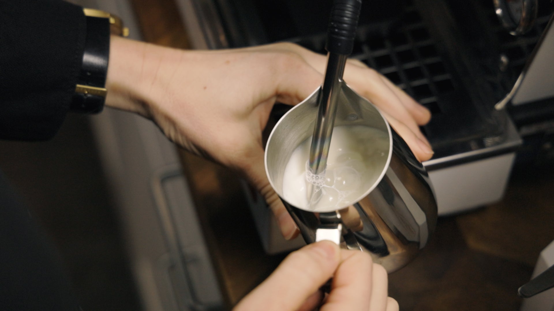 Hands frothing milk in a stainless steel pitcher under an espresso machine steam wand.