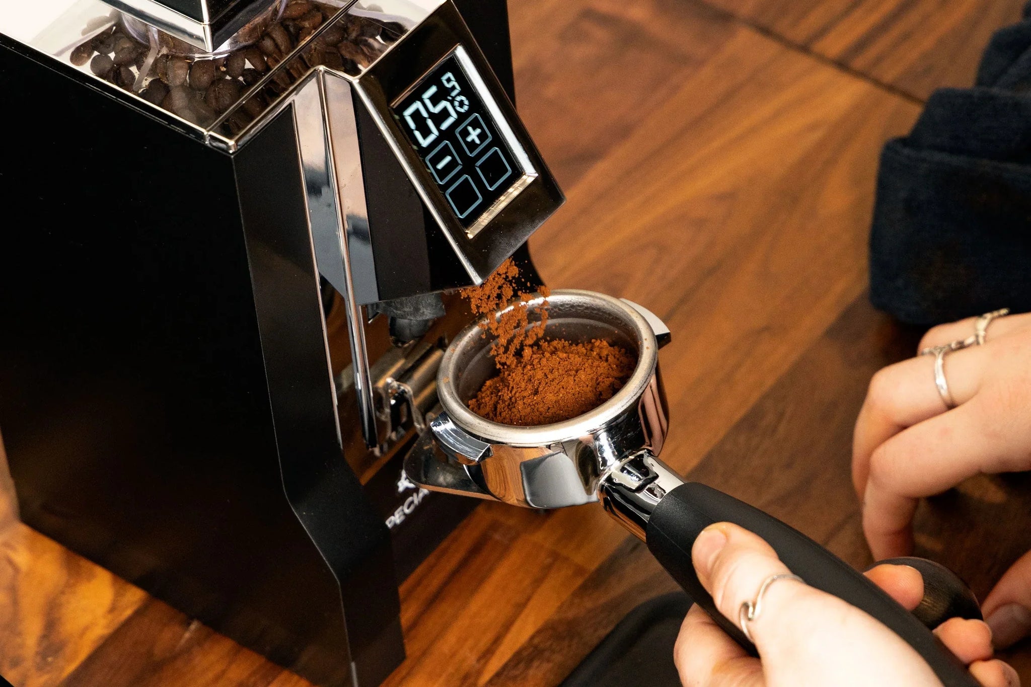 Close up of a grinder dispensing espresso grounds into a metal portafilter with coffee beans visible in the hopper.