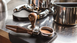 Close up of a wooden espresso tamper resting on the portafilter with a shiny espresso machine in the background