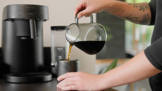 Tattooed arm pours coffee from a glass carafe into a metal mug beside a black coffee maker.