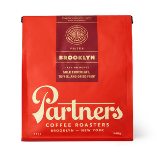 Red Brooklyn coffee bag beside a glass carafe pouring coffee into a white cup.