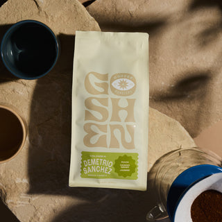 Colombia Demetrio Sanchez Gesha coffee bag placed on a sandy surface beside ceramic mugs