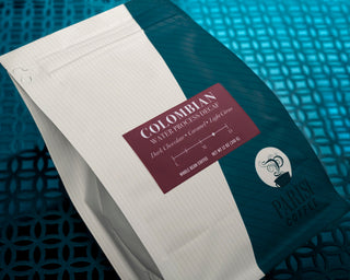 Colombian Decaf coffee bag beside a notebook on a wooden table with a cup of coffee and biscotti
