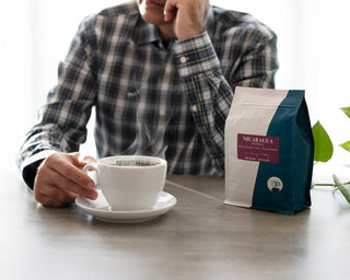 Man in plaid shirt sits at a table with a steaming coffee cup and a Nicaragua Jinotega bag.