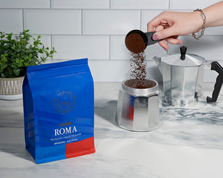 Hands pour ground coffee from a scoop into a moka pot beside a blue Roma coffee bag on a kitchen counter