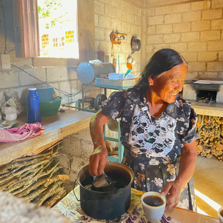 Mazatec woman in a floral dress smiles in a sunlit rustic workshop with coffee tools and exposed block walls.