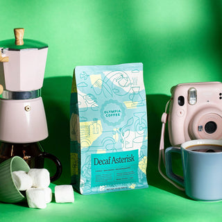 Decaf Asterisk bag with teal artwork stands upright on a green studio backdrop beside a white coffee grinder.
