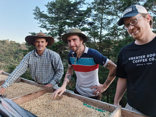 Three Greater Goods crew members wearing wide brim hats inspect a wooden drying bed filled with coffee beans outdoors