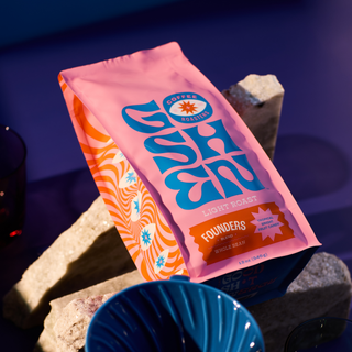 Pink Founders Blend bag from Goshen Coffee Roasters with blue logo, set against rocks and a dark background.