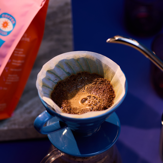 Blue coffee mug with frothy brew beside pink Founders packaging in the background.