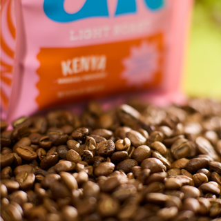 Close-up of roasted coffee beans with a pink Kenya Mbegu Ndogo Whole Bean bag in the background