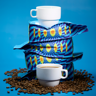 White ceramic cup sits atop a blue patterned Old School Tattoo coffee bag with coffee beans scattered on a blue surface