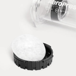 Round white AeroPress paper filter resting on the black filter cap with the transparent AeroPress chamber in the background
