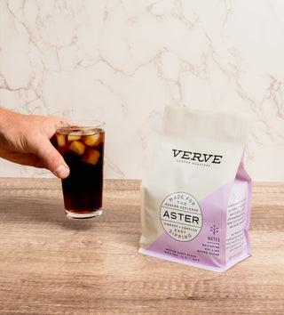 Hand holding a glass of iced coffee beside a Verve Aster coffee bag on a marble countertop.