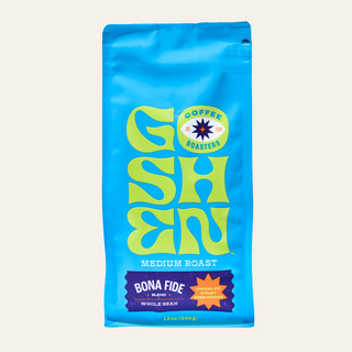 Blue Bona Fide bag on a blue surface beside a white cup of coffee.