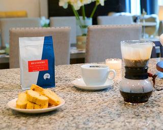 Bravissimo coffee bag on a granite countertop beside a white cup and pastries in a cafe setting.