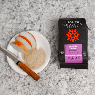 Bolivian Caranavi coffee bag shown beside a plate with pastry and a cinnamon stick on a speckled countertop.
