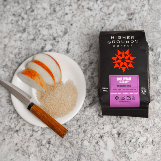 Bolivian Caranavi coffee bag shown beside a plate with pastry and a cinnamon stick on a speckled countertop.