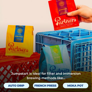 Jumpstart yellow coffee bag from Partners Roasters displayed on a red crate with a hand reaching a red bag.