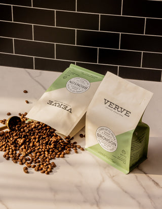 Bronson coffee bag on a green surface with scattered chocolate chunks and a jar nearby