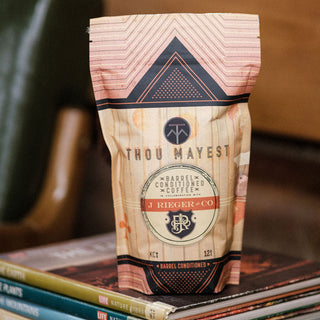 Thou Mayest barrel conditioned coffee bag with pink geometric top and crest logo resting on magazines.