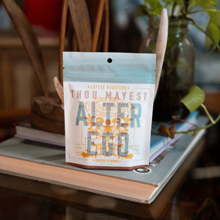 A person holds a Thou Mayest coffee bag with illustrated label and visible tag against a plant filled background.