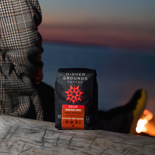 Person in a plaid shirt at a table with a decaf bag from Higher Grounds in focus against a sunset