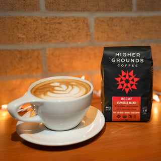 Latte art in a white cup placed beside a Higher Grounds Decaf bag against a brick wall