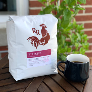 White coffee bag with a red rooster logo sits on a wooden table beside a cup, with green plants in the background.
