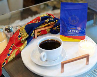 Bravissimo coffee bag on a cafe table beside a white cup of coffee and a colorful scarf in the background.